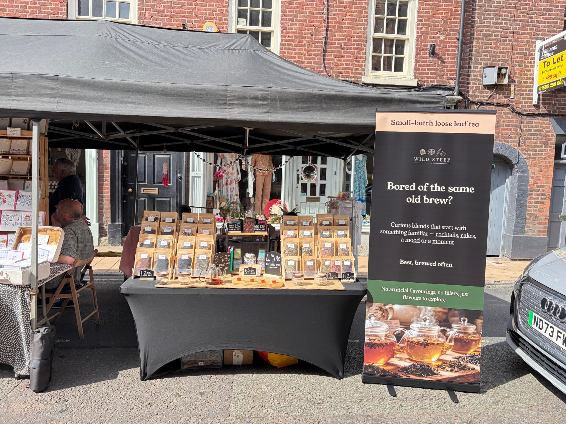 The Wild Steep market stall on a sunny high street, kraft pouches of loose leaf tea racked on a table beside a branded banner.